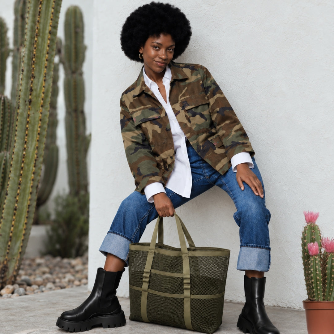 Woman wearing camo jacket and carrying a green tactical mesh tote leaning against a wall with cactus next to her. 