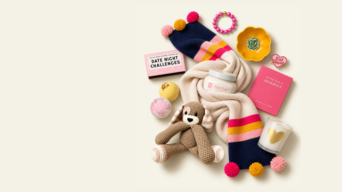A styled flat-lay of multiple cozy products on a warm off-white background, including a knitted bunny toy, a cream knit scarf with pink and navy striped ends, a pink bath bomb, a pink date-night challenge card, and a jar labeled nectar. Scarf, book, toy, bath bombs, candle, bracelet, and fashion-forward gifts that give back and donate to charity with purchase at The Kindness Cause