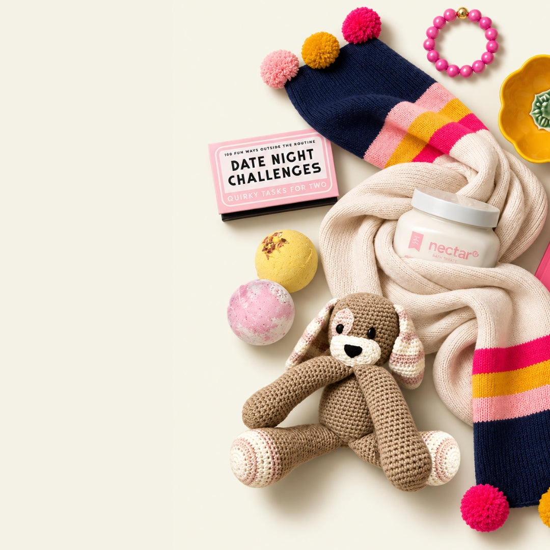 A styled flat-lay of multiple cozy products on a warm off-white background, including a knitted bunny toy, a cream knit scarf with pink and navy striped ends, a pink bath bomb, a pink date-night challenge card, and a jar labeled nectar. Scarf, book, toy, bath bombs, candle, bracelet, and fashion-forward gifts that give back and donate to charity with purchase at The Kindness Cause