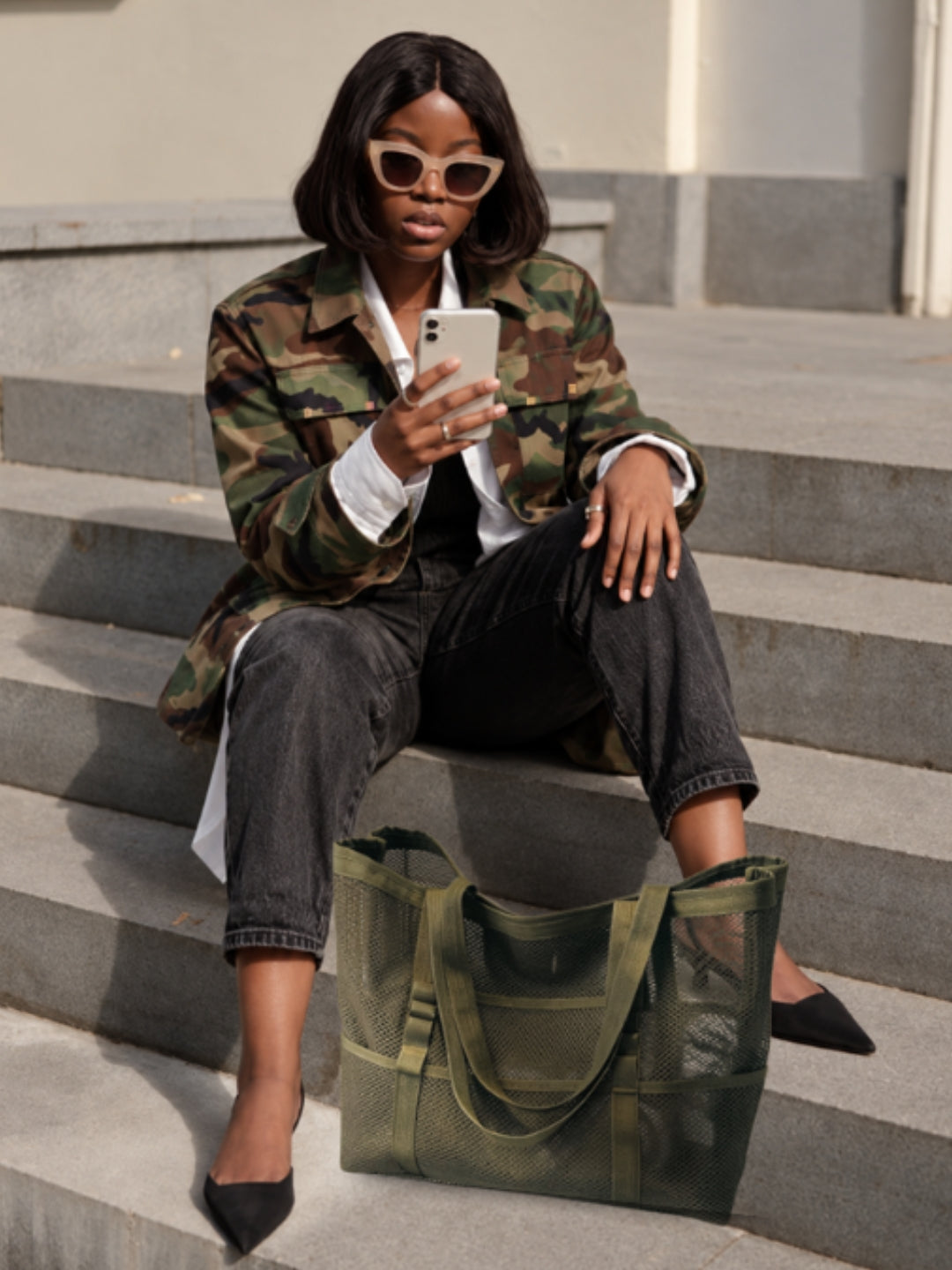 Woman wearing a camo jacket on her phone with green tote bag.  Shop Camo Gifts That Give Back. 